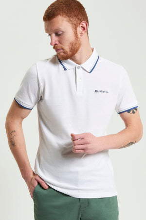 A man with short red hair and a beard wears a white Ben Sherman Signature Pique Polo with blue trim and green pants. Made from organic cotton pique, he stands with one hand in his pocket, looking down to his left.