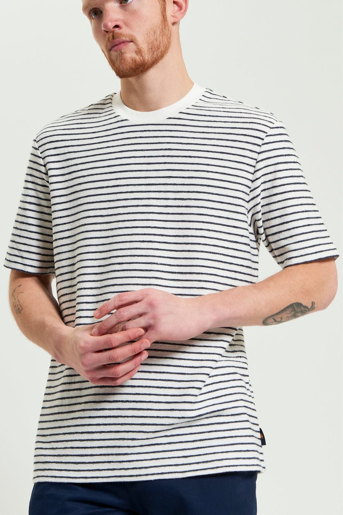 A man with a short red beard wears a Ben Sherman Textured Stripe Tee, paired with navy pants. He stands with hands clasped, displaying tattoos on his left forearm.