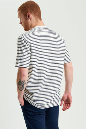 A man with short red hair and a beard, wearing a Ben Sherman Textured Stripe Tee and navy pants, stands with his back to the camera. A heart-shaped tattoo is visible on his right forearm.