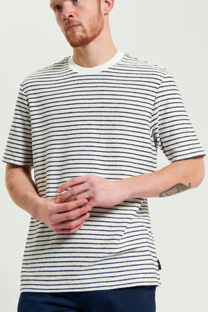 A man with a short red beard wears a Ben Sherman Textured Stripe Tee, paired with navy pants. He stands with hands clasped, displaying tattoos on his left forearm.