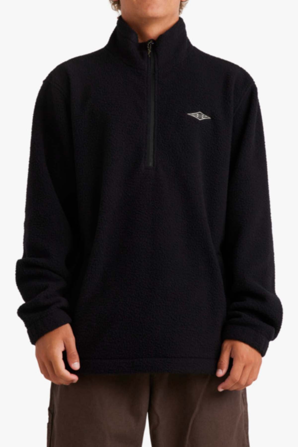 Wearing the Billabong A/Div Boundary Mock Neck Sweatshirt, a black fleece pullover made of recycled polyester with a half-zip and an embroidered chest logo, a person stands relaxed in brown pants. The image is cropped at the neck and mid-thigh.