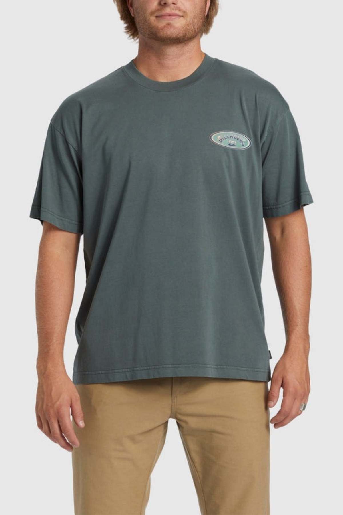 A person standing and wearing a Billabong Arch Wave T-Shirt in dark green, made from heavyweight cotton fabric, with a small emblem on the left chest, and paired with khaki pants. The background is plain and light-colored.