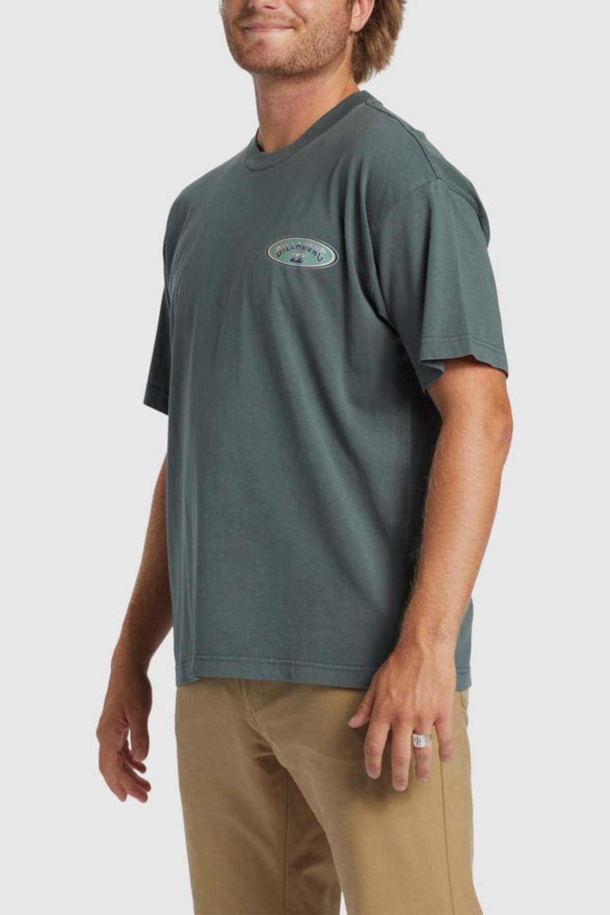 A person wearing a Billabong Arch Wave T-Shirt in dark green, featuring a small oval logo on the chest, paired with khaki pants. The individual has light-colored hair and is smiling. The heavyweight cotton fabric of the OG fit shirt stands out against the plain, light-colored background.