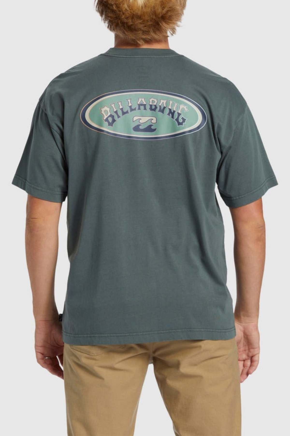 A person with short, light brown hair is wearing a greenish-gray Billabong Arch Wave T-Shirt made from heavyweight cotton fabric. The shirt features an oval logo on the back that says "Billabong" and showcases a wave graphic. This person is also sporting beige pants and is facing away from the camera.