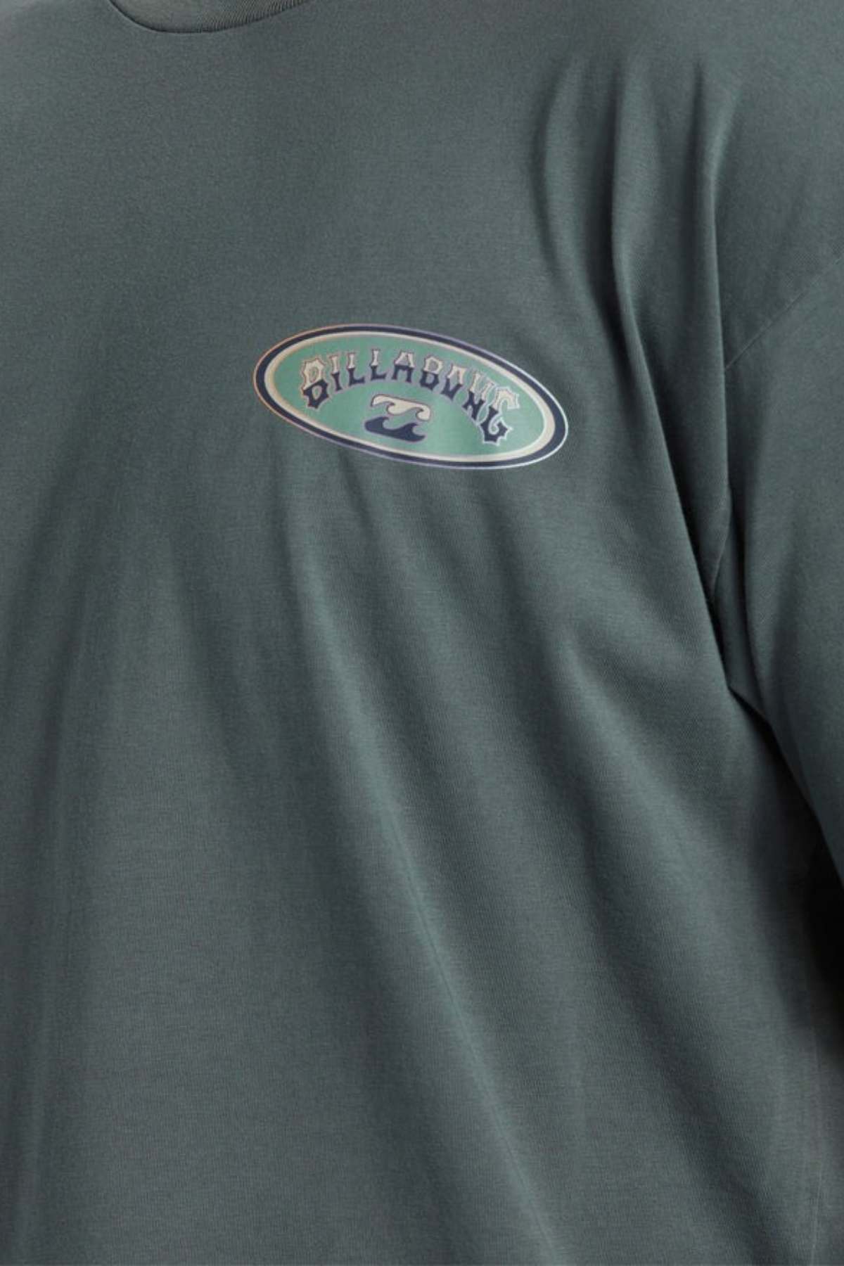 Close-up of a person wearing a dark green Billabong Arch Wave T-Shirt in the OG fit. This heavyweight cotton fabric shirt showcases an oval logo on the upper left side, featuring the text "Billabong" alongside a stylized wave design.