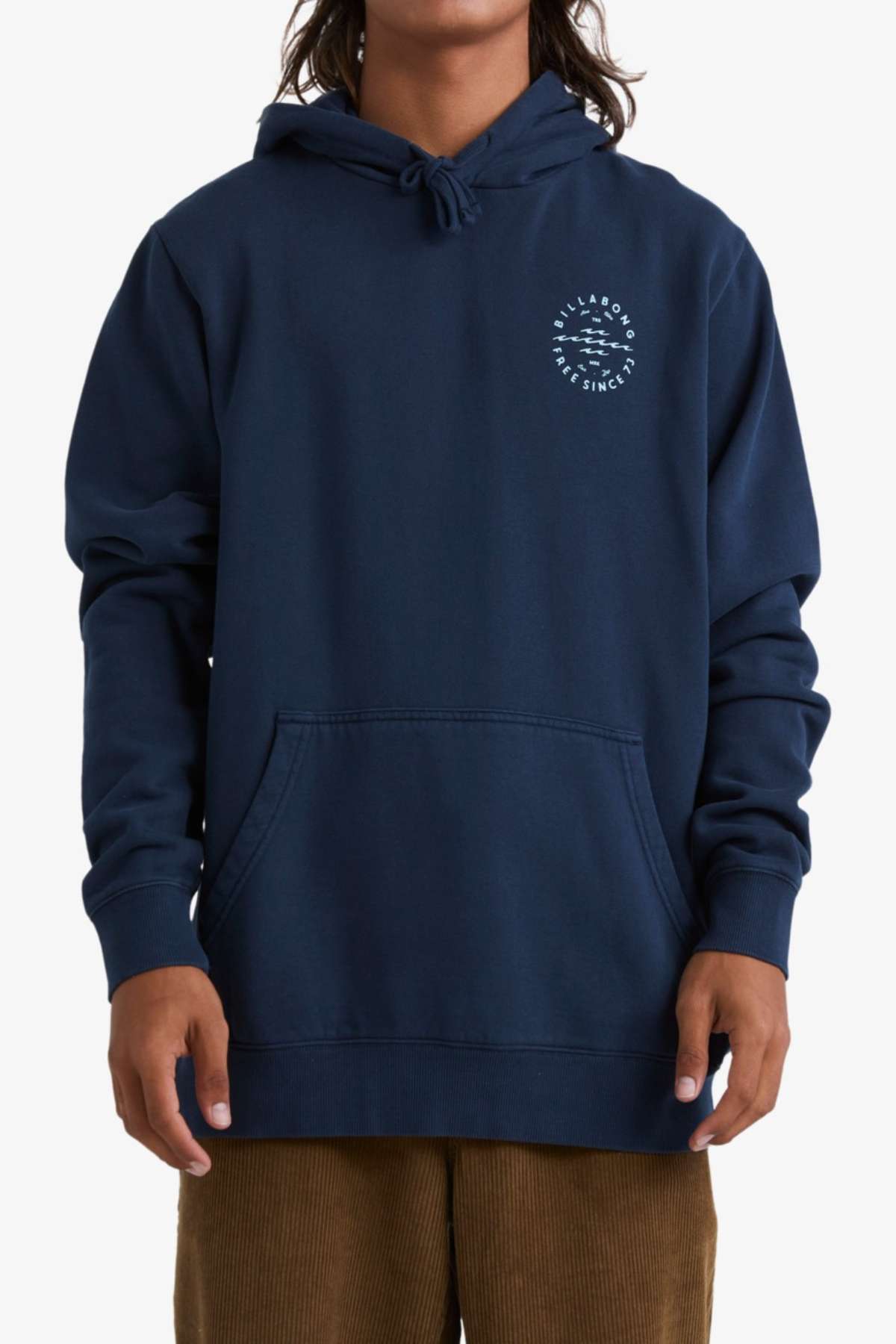 Someone with long hair is sporting a navy blue Billabong Big Wave Daz Pop Hood, which features a small, circular logo on the left chest. The cozy half brushed fleece hoodie includes a front pocket and a pulled-up hood. Perfect for casual outings, they are also wearing brown corduroy pants.