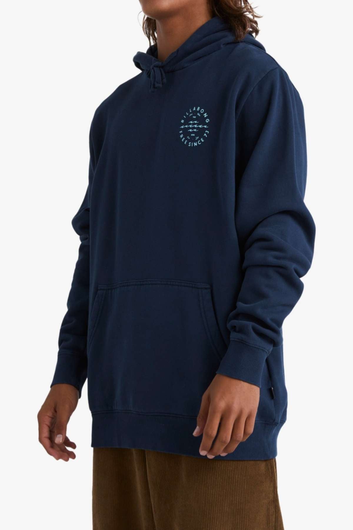 A person wearing the Billabong Big Wave Daz Pop Hood, a dark blue hooded sweatshirt crafted from cozy half brushed fleece and featuring a small circular logo on the left chest. The person has long hair and is standing with their hands relaxed by their sides, also dressed in brown pants, against a plain white background.