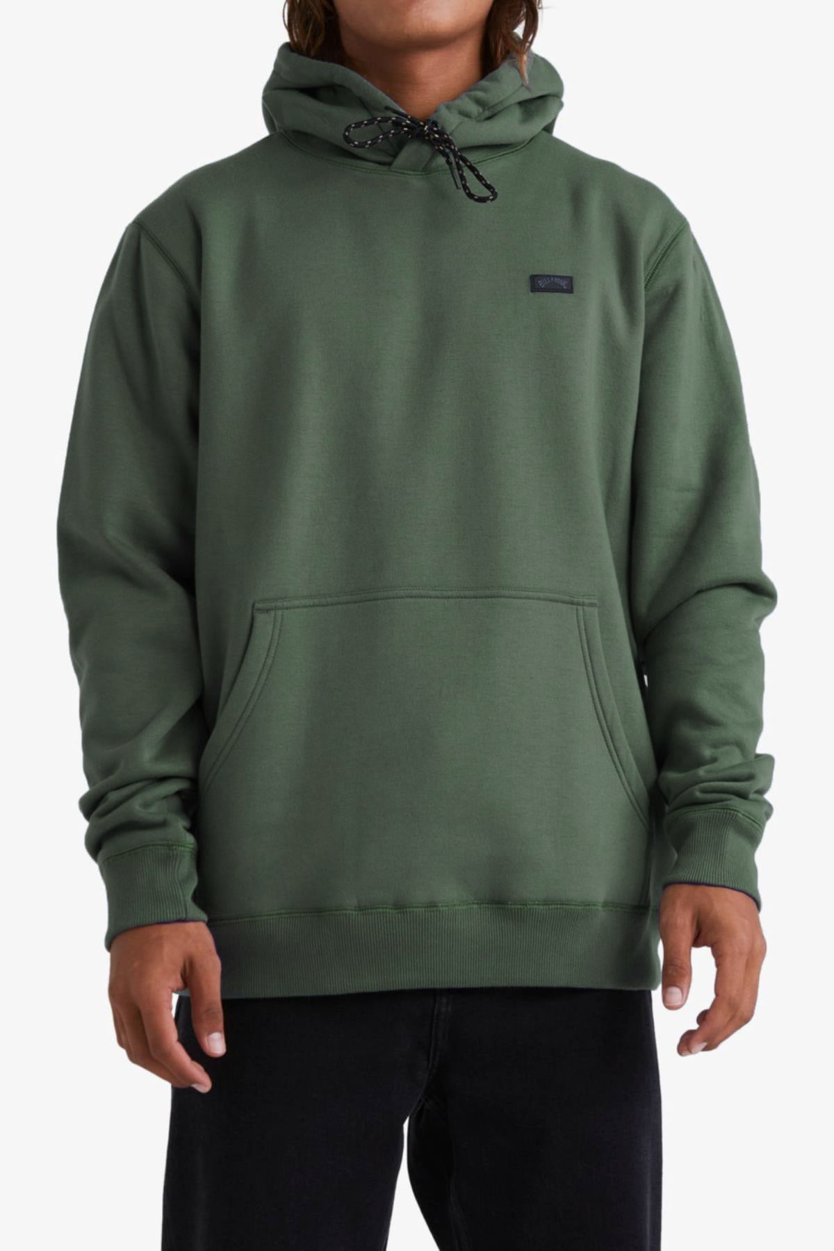 A person in a forest green Billabong Shoreline Pullover with cozy fleece lining and black drawstrings, paired with black pants, stands against a white background.