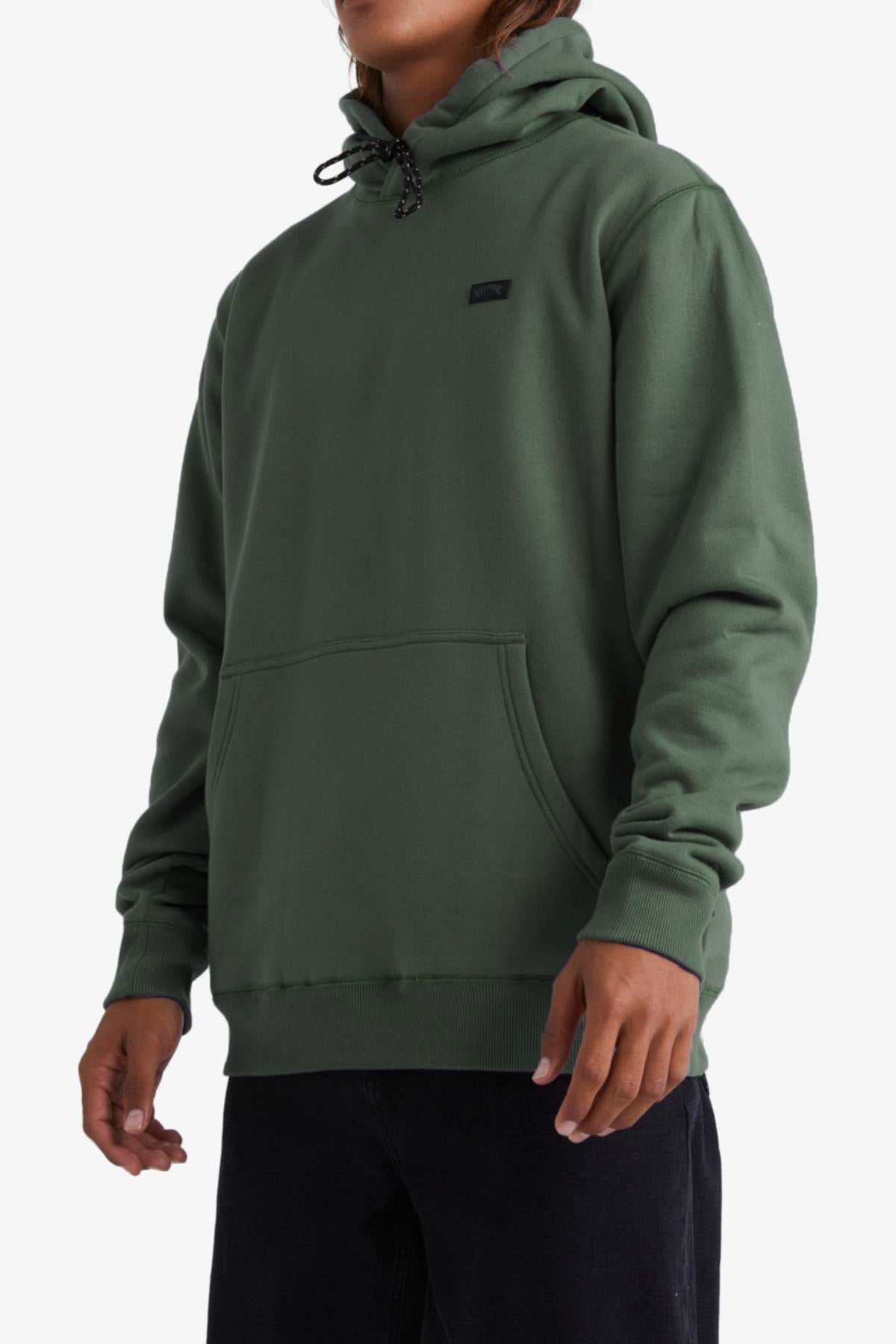 A person wearing the Billabong Shoreline Pullover—a green fleece-lined hoodie with a front pocket and black drawstrings—paired with black pants, stands against a plain white background.