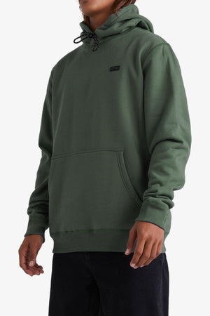A person wearing the Billabong Shoreline Pullover—a green fleece-lined hoodie with a front pocket and black drawstrings—paired with black pants, stands against a plain white background.