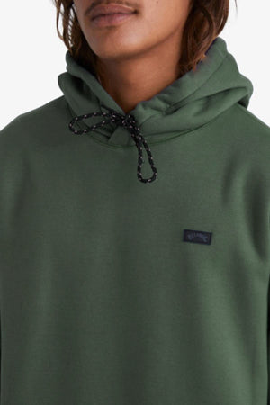 A person with a mustache and long hair wears a forest green Billabong Shoreline Pullover featuring black drawstrings, a small black chest patch, and visible cozy fleece lining.