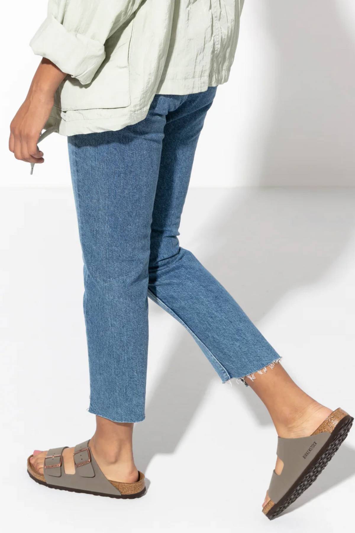 A person is shown from the waist down wearing a light green shirt, blue cropped jeans, and taupe Birkenstock Arizona sandals with adjustable buckles and a contoured footbed, walking on a white background.