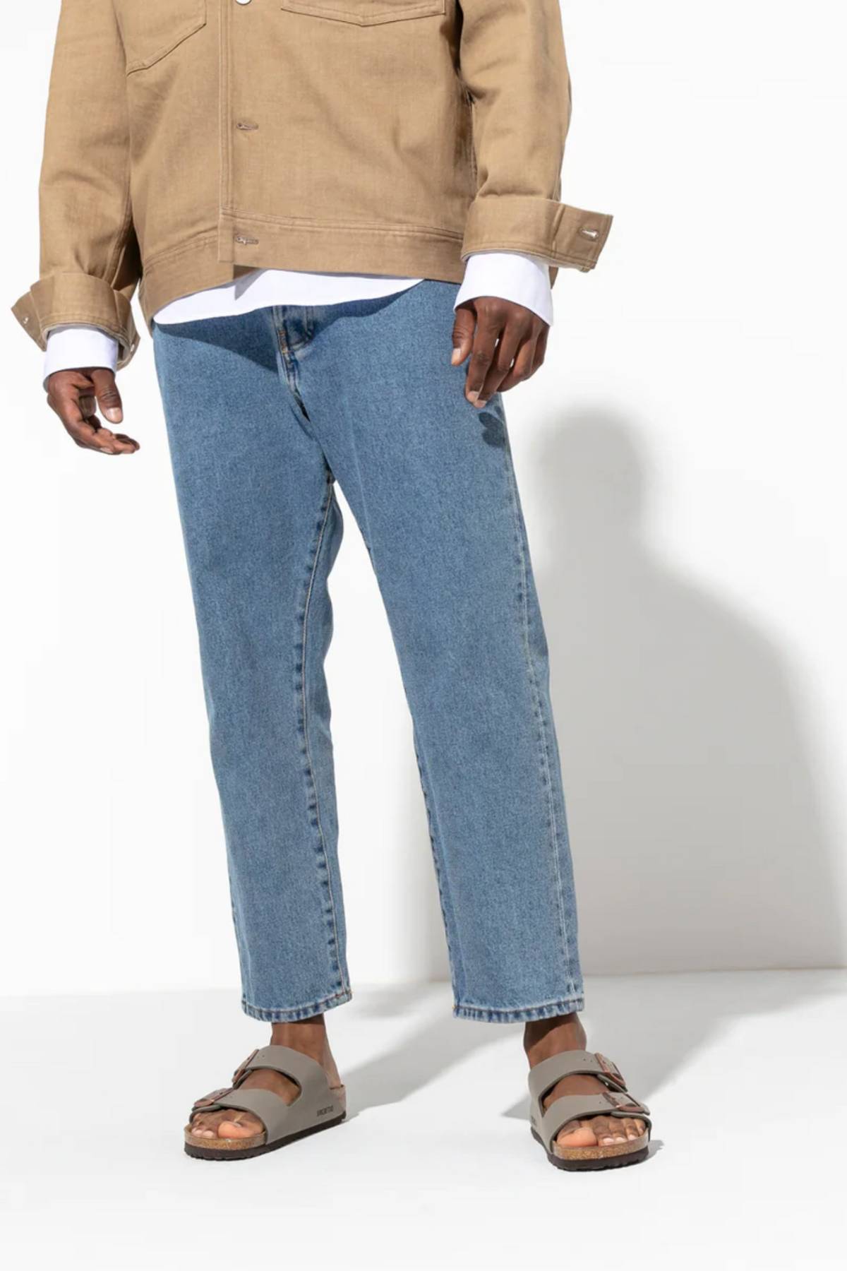 A person wearing a tan jacket, white shirt, blue jeans, and gray Birkenstock Arizona sandals stands against a plain white background. The image is cropped from the shoulders down to highlight the Birkenstock Arizona footwear.