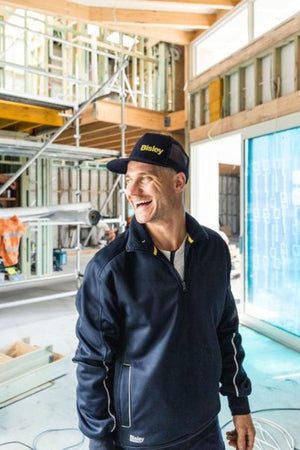A smiling man in a navy Bisley 1/4 Zip Sherpa Lined Fleece and cap stands inside a partially constructed building with exposed framing and large windows.