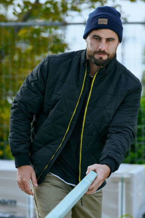 A bearded man in a navy beanie, beige pants, and a Bisley Quilted Bomber Jacket holds a long wooden plank outdoors, with trees and a metal fence behind him.