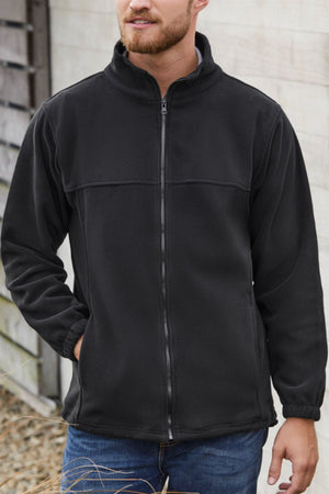 A man wearing the Biz Collection Plain Micro Fleece Jacket in black (100% polyester, full zip), with his left hand in his pocket, stands casually outdoors in blue jeans against a wooden wall.