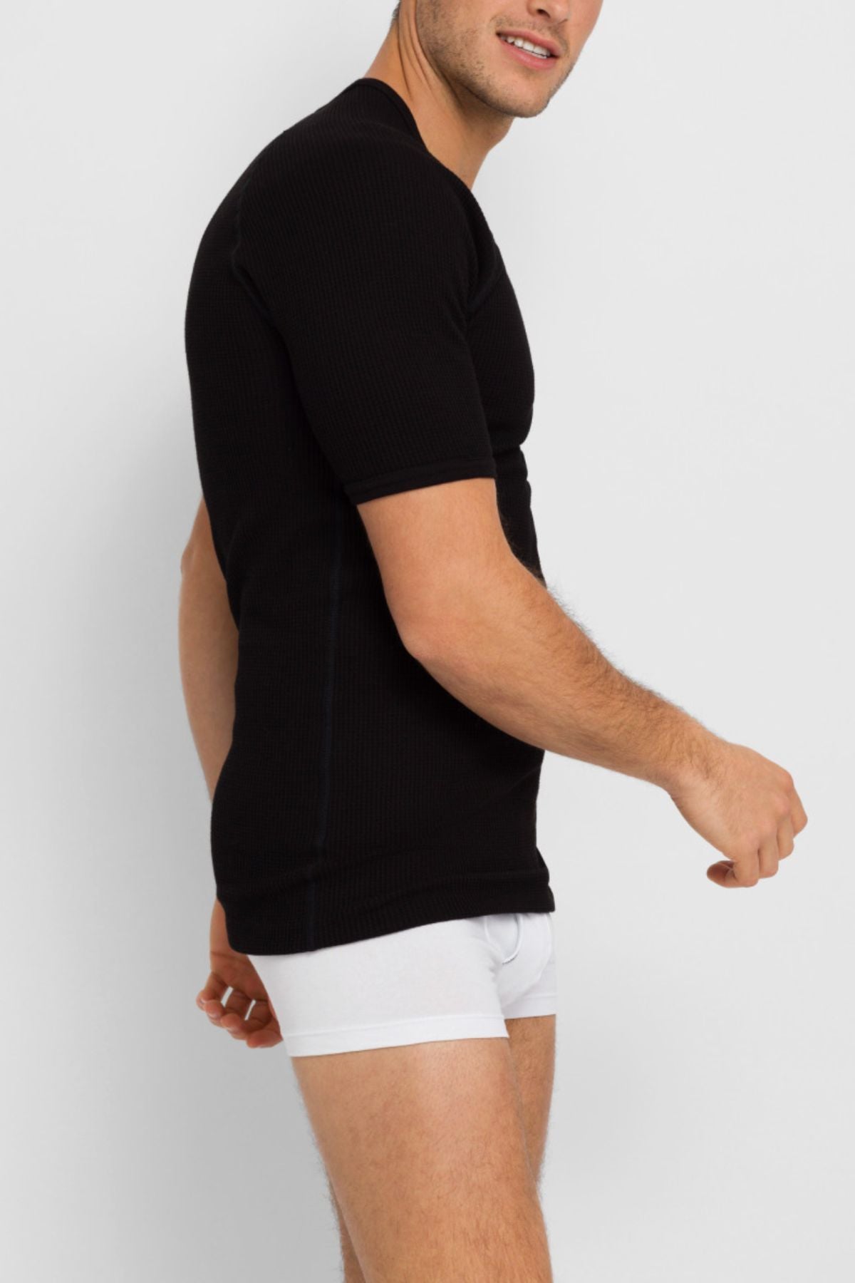 A man wears a fitted Bonds Aircel Short Sleeve Tee in black with white boxer briefs, standing against a plain light background, looking slightly over his shoulder and smiling.