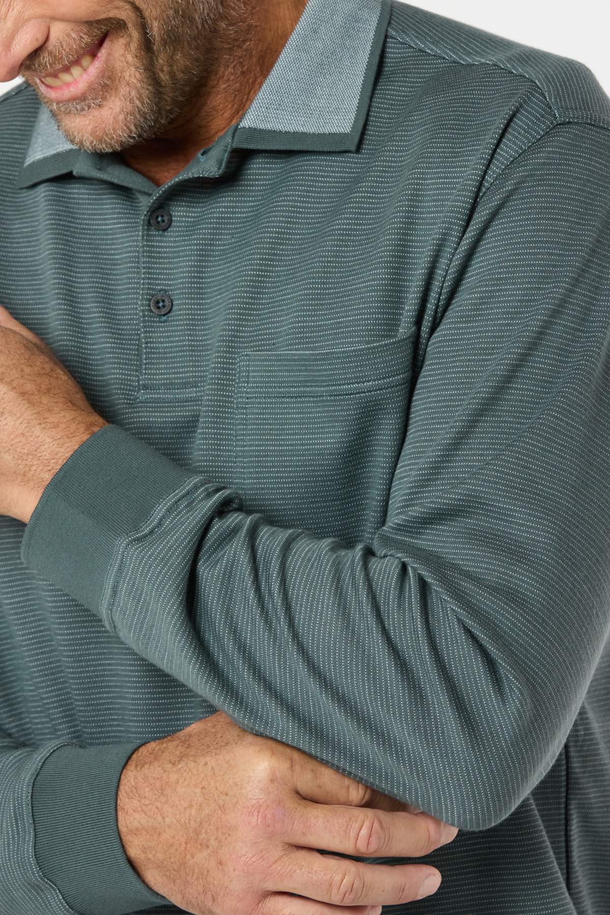 A smiling man in a Breakaway Avantee Micro Prima Polo, featuring a textured teal finish, rib knit cuff, chest pocket, and button collar, crosses his arms with only part of his face and upper body visible against a white background.