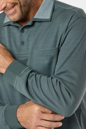 A smiling man in a Breakaway Avantee Micro Prima Polo, featuring a textured teal finish, rib knit cuff, chest pocket, and button collar, crosses his arms with only part of his face and upper body visible against a white background.