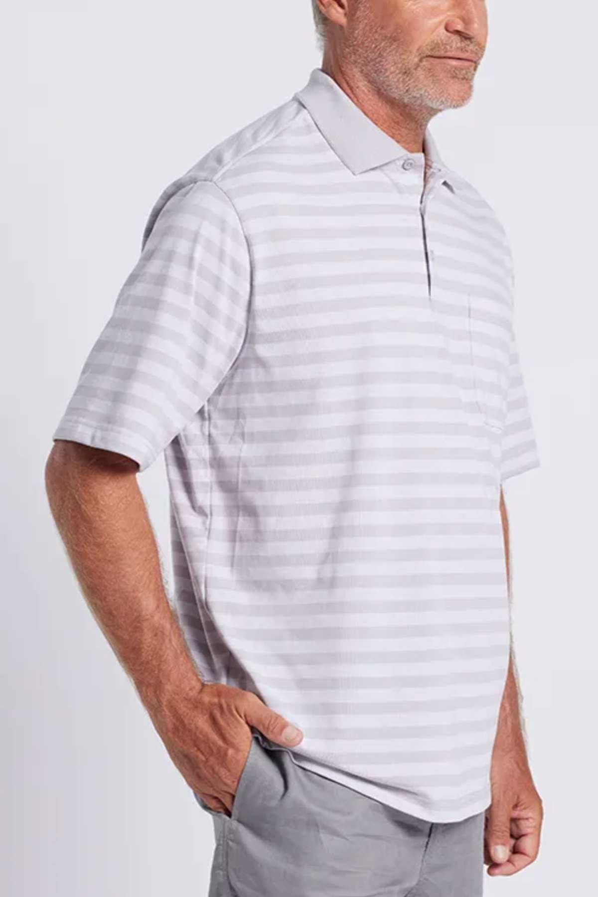 A man wearing a Breakaway Brady Kinnersly Polo, which is stylish and comfortable, stands with one hand in his pocket. The light gray and white striped shirt showcases a contrast rib knit collar and short sleeves. He pairs it effortlessly with light gray pants against a plain, light-colored background.