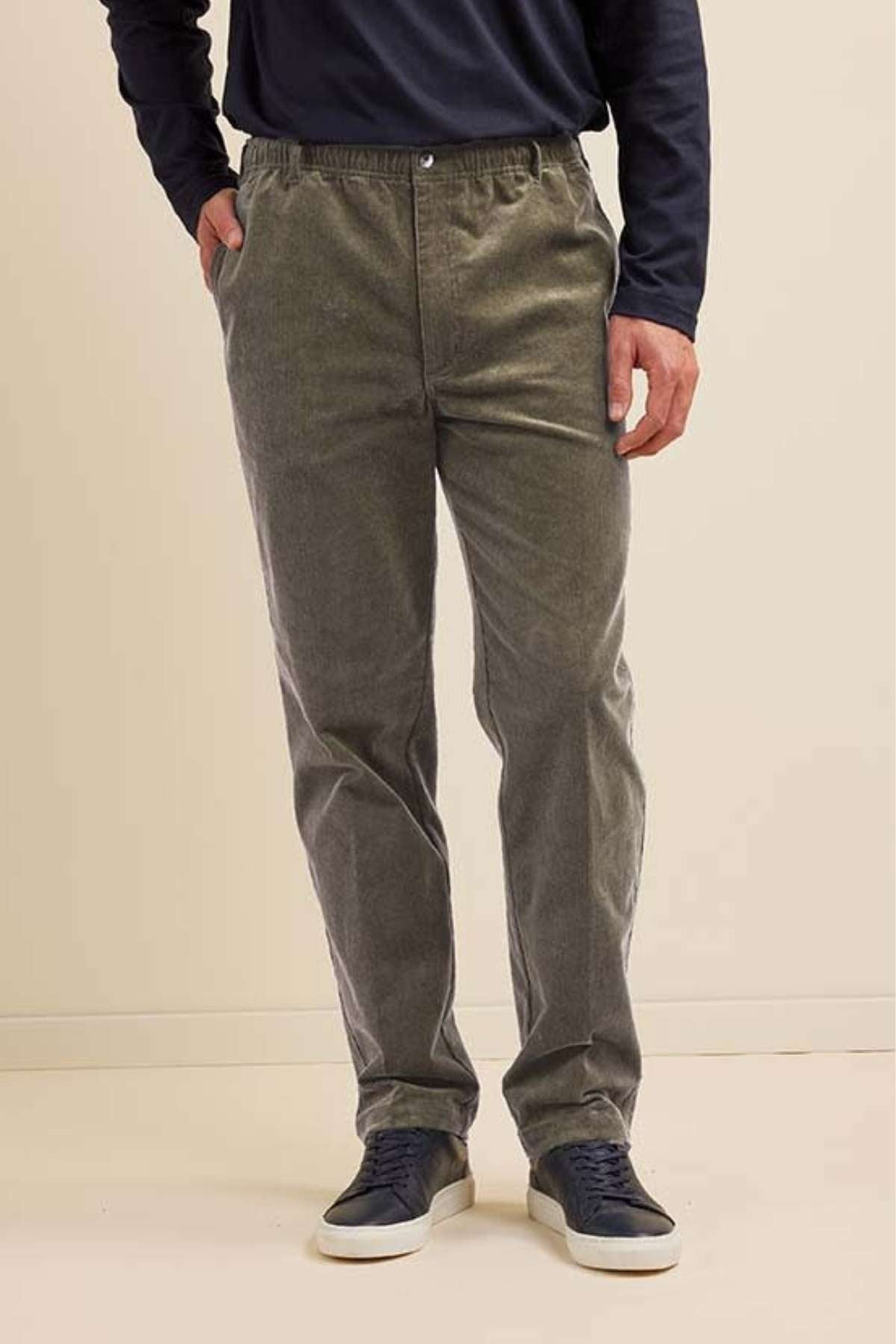A person wears Breakaway Cordeaux Pant with an elastic waist and stretch fabric, paired with a dark long-sleeve shirt and black sneakers, standing with one hand in their pocket against a plain light background.