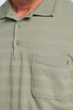 A close-up of a person wearing a light green Breakaway Emil Polo, showcasing its self-jacquard stripe pattern. This 100% cotton shirt features a front chest pocket with a subtle embroidered detail and has two buttons undone at the collar.