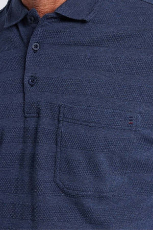 A close-up shot showcases a person donning the Breakaway Emil Polo in dark blue, distinguished by its elegant knit collar and chest pocket. Crafted from 100% cotton, this textured polo features a three-button placket and subtle horizontal stripes. The upper torso is prominently visible.