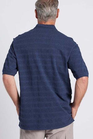 A man with short, gray hair stands with his back to the camera, dressed in a dark blue Breakaway Emil Polo that boasts an elegant knit collar, paired with light gray pants. Made from 100% cotton fabric, the polo ensures comfort. The background is plain white.