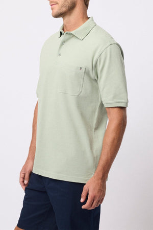 A man wears the Breakaway Fraser Cotton Pique Polo in light green with a chest pocket, paired with dark navy shorts, standing against a plain light background.