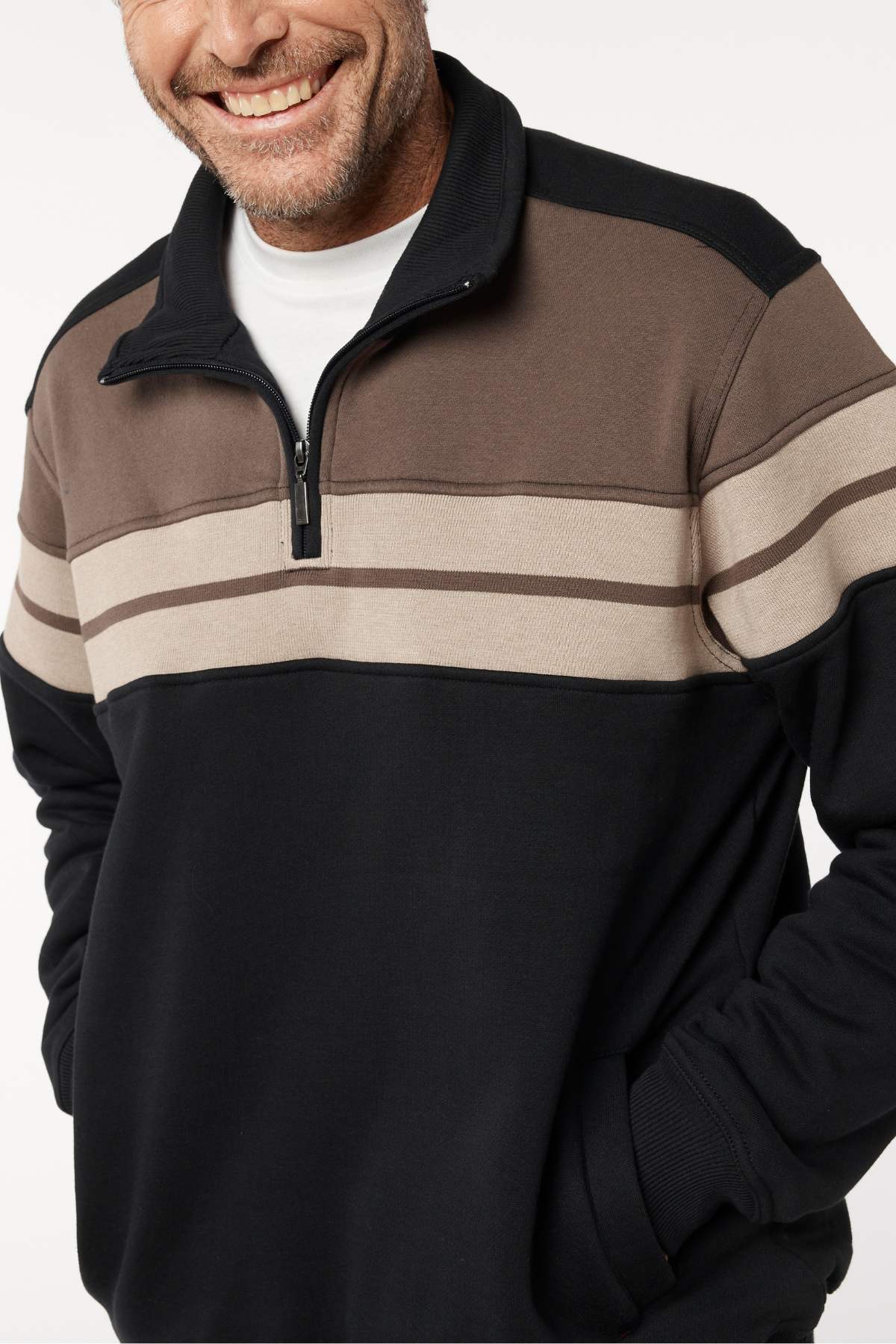 A man smiles with his hands in his pockets, wearing a Breakaway Henrich Snowy Mt Fleece Half Zip with wide black and brown stripes and beige accents over a white undershirt. Only his upper body is visible.