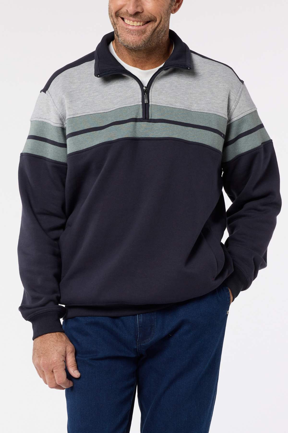 A man wears a Breakaway Henrich Snowy Mt Fleece Half Zip in navy blue and gray with green and gray chest stripes, paired with blue jeans. He stands smiling with one hand in his pocket.