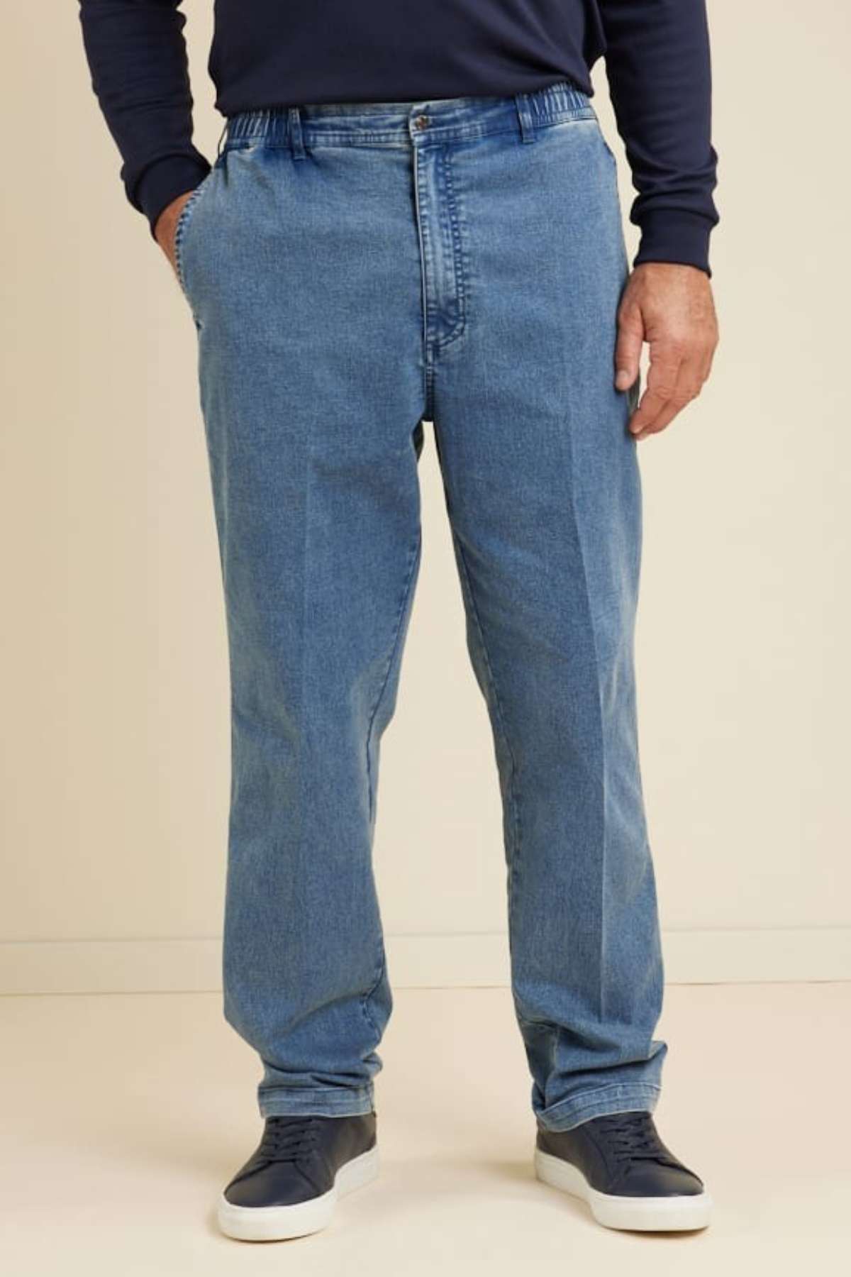 Someone stands against a plain light background wearing Breakaway Intense Denim Jeans with an elastic waist, a dark long-sleeve shirt, and dark sneakers, their hands in their pockets.