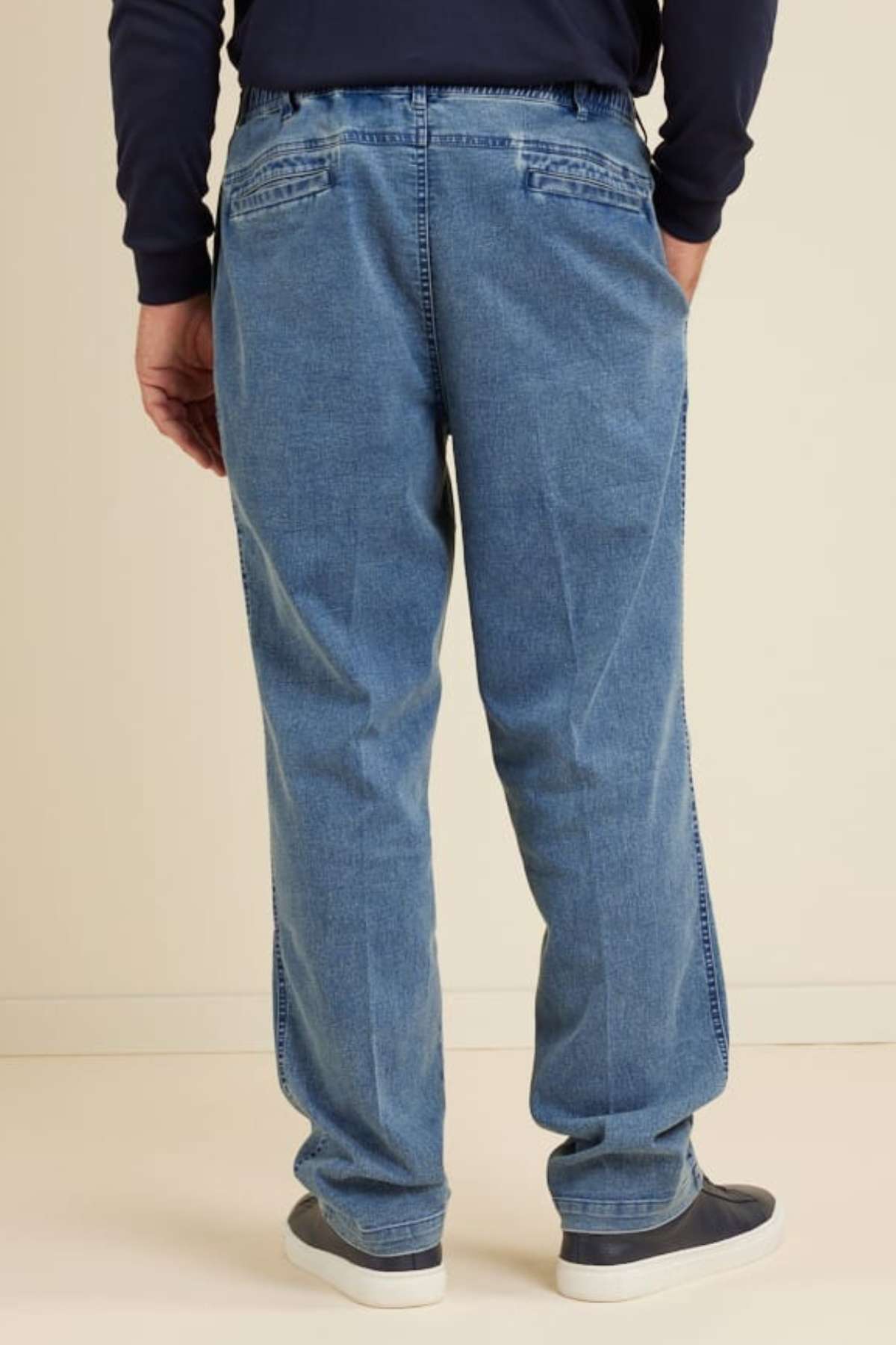 Wearing the Breakaway Intense Denim Jean in light blue with an elastic waist and straight leg, a person stands facing away in a navy long-sleeve shirt and dark slip-on shoes against a plain, light-colored background.