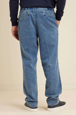 Wearing the Breakaway Intense Denim Jean in light blue with an elastic waist and straight leg, a person stands facing away in a navy long-sleeve shirt and dark slip-on shoes against a plain, light-colored background.