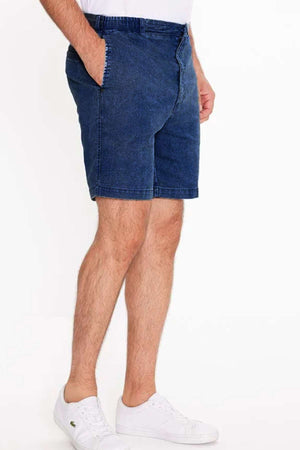 A man stands with hands in pockets, wearing Breakaway Intense Denim Shorts in garment-dyed mid-blue and white sneakers against a plain white background.