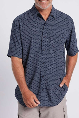 A person with a light beard and mustache is smiling, wearing a short-sleeve Breakaway Jac Bamboo Shirt in dark blue paired with beige cargo shorts. Their right hand rests in their shorts pocket, while the left hand hangs relaxed by their side against a plain white background.