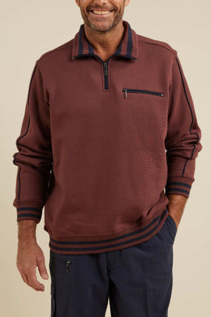 A smiling man stands with one hand in his pocket, wearing the Breakaway Kyler Snowy Mt Fleece Half Zip, a maroon long-sleeve quarter-zip sweatshirt with a striped collar and cuffs. This soft and warm fleece features a vertical zipper pocket on the chest. He pairs it stylishly this winter with navy blue pants.