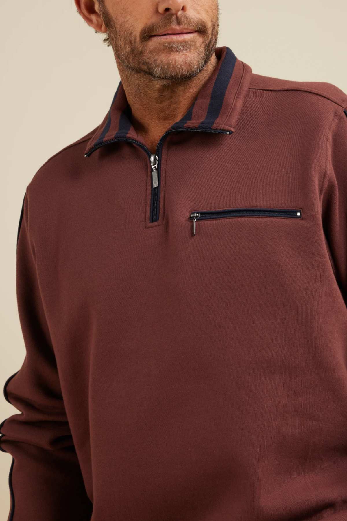 A man is shown from the shoulders up, wearing a Breakaway Kyler Snowy Mt Fleece Half Zip in brown with a navy blue stripe on the collar and a zippered chest pocket. He has light facial hair and is looking away from the camera. This stylish winter piece boasts soft and warm fleece, perfect for chilly days. The background is a plain, neutral color.