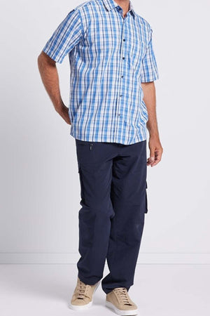 A person standing against a plain background, exuding effortless elegance in a Breakaway Ora Bamboo Blend Shirt with short sleeves and blue and white plaid pattern, paired with dark blue cargo pants and beige sneakers. Their arms rest casually by their sides.
