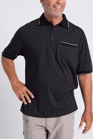 A smiling person stands against a plain background, wearing a black short-sleeved Breakaway Pete Kinnersly Polo featuring a subtle geometric print and light-colored accents on the collar, pocket, and sleeve edges. With their right hand on their hip, the person complements the outfit with light gray pants.