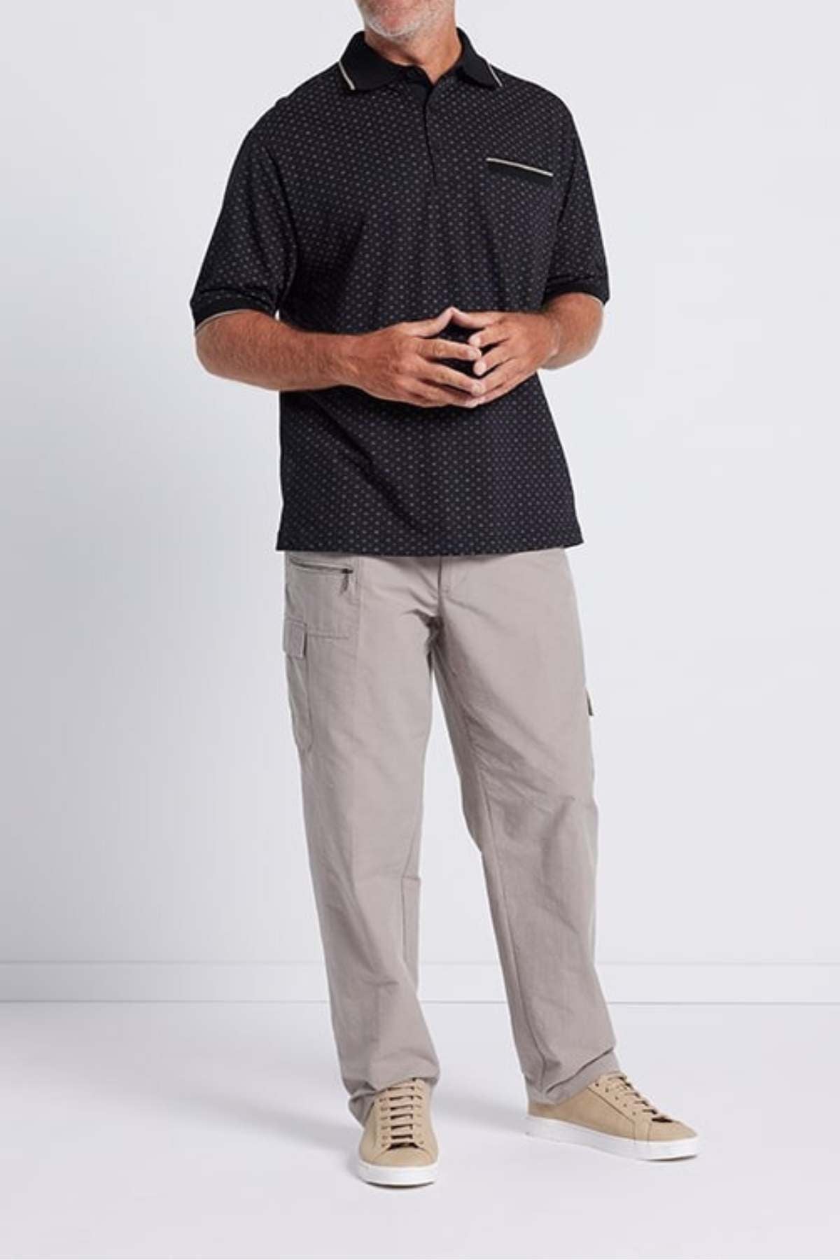 A person stands against a plain background, wearing the Breakaway Pete Kinnersly Polo featuring a dark geometric print and short sleeves along with a breast pocket, paired with light gray cargo pants and beige sneakers. They are holding their hands together in front of their body, with their face not visible.