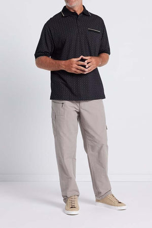 A person stands against a plain background, wearing the Breakaway Pete Kinnersly Polo featuring a dark geometric print and short sleeves along with a breast pocket, paired with light gray cargo pants and beige sneakers. They are holding their hands together in front of their body, with their face not visible.