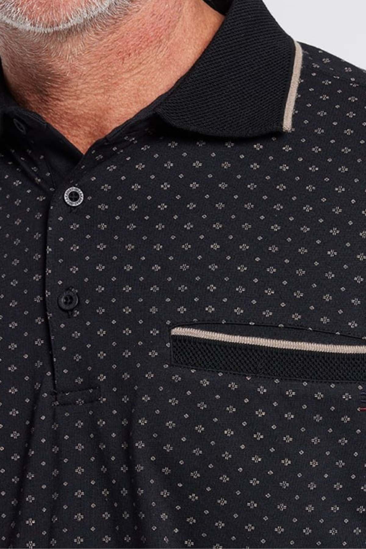 Close-up of a man wearing a Breakaway Pete Kinnersly Polo adorned with small white flower patterns. The shirt features a black collar with beige trim, black buttons, and a pocket on the left side with an opening bordered in beige. The man's beard and part of his neck are visible.