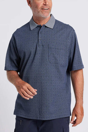 A man with short gray hair and a beard wears the Breakaway Rye Micro Prima Polo in Denim Marle with a birdseye check, light gray collar, and chest pocket. He smiles and looks left against a plain light gray background.
