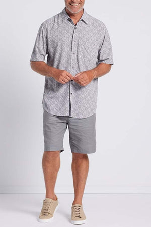 A man stands against a plain background wearing the Breakaway Shaw Bamboo Shirt with a sophisticated leaf print, gray shorts with a smart pocket, and beige sneakers. He smiles and holds his hands together.