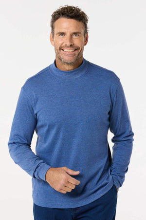 A smiling man with curly hair and a beard poses against a plain white background, wearing the Breakaway Skivvy—a long-sleeve blue shirt crafted from a polyester-cotton blend. One hand rests in his pocket, creating a timeless winter look.