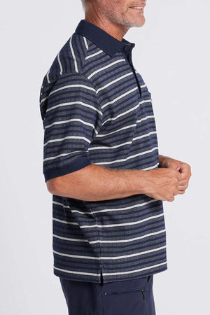 A man, standing sideways, is wearing a Breakaway Turner Micro Prima Polo that features a striped pattern in multiple shades of blue, gray, and white. He has a short beard and his hands are clasped in front of him. The background is plain and light gray.