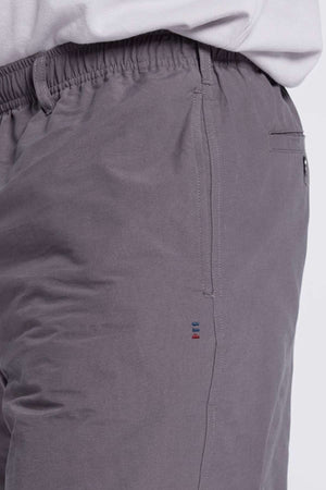 Close-up of a person wearing gray Breakaway Woodbury Short with an elastic and adjustable waistband and a side pocket. Near the pocket seam, there is a small embroidered design featuring three vertical lines in different colors. The edge of a light-colored shirt is partially visible above the waistband.