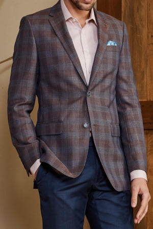 A person wears the Bruton Abram Linen Blazer in brown plaid, featuring a wool/linen blend and pocket square, paired with a pink shirt and blue trousers. They stand against wooden panels with one hand in their pocket, showcasing a contemporary fit.