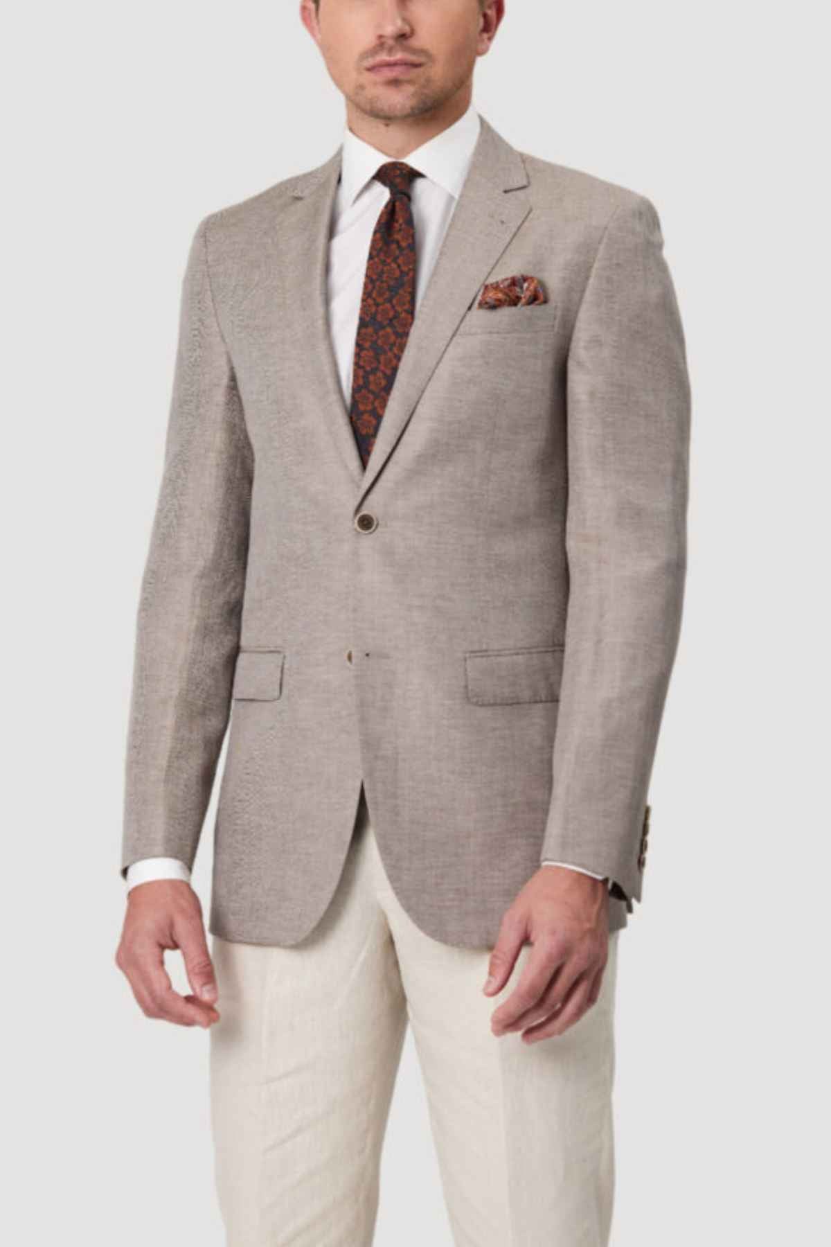 A man showcases luxury fashion by wearing the Bruton Abram Linen Blazer in light gray, paired with a dark patterned tie and matching pocket square. The ensemble is perfectly completed with crisp white trousers, striking a balance between sophistication and style.